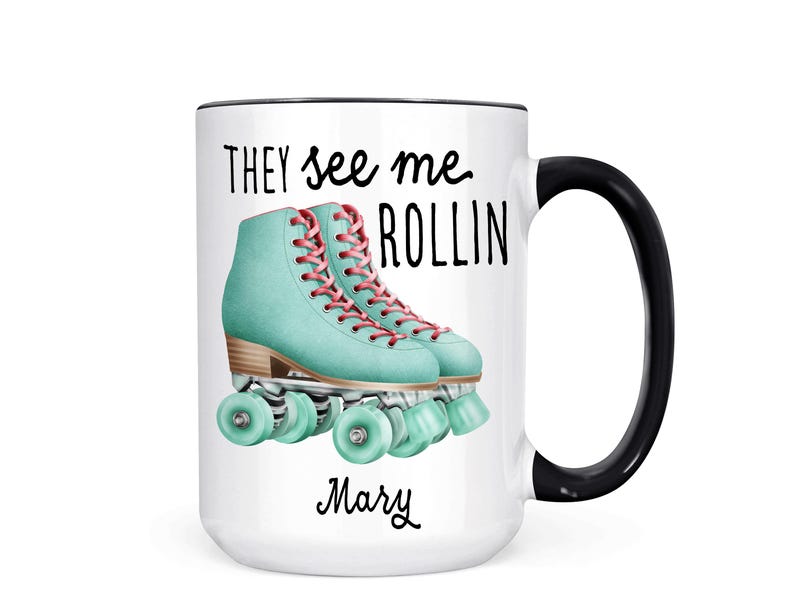 Op de afbeelding: Witte keramische koffiemok met een zwarte rand en handvat. De mok heeft een afbeelding van twee turquoise rollerskates met roze veters en de tekst "They see me rollin'" en de naam "Mary" onderaan.