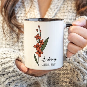 May include: White ceramic mug with a black rim and handle. The mug features a line drawing of a gladiolus flower in orange and green. The text "Aubrey, Gladiolus - August" is printed on the mug.