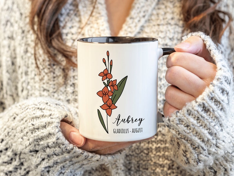 May include: White ceramic mug with a black rim and handle. The mug features a line drawing of a gladiolus flower in orange and green. The text "Aubrey, Gladiolus - August" is printed on the mug.