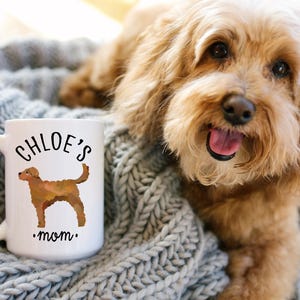 May include: White ceramic mug with the text "CHLOE'S mom" and a brown dog illustration. The mug is on a gray knitted blanket, with a light brown dog in the background.