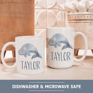May include: Two white ceramic mugs with a blue whale design and the name "Taylor" printed on each. The mugs are dishwasher and microwave safe.