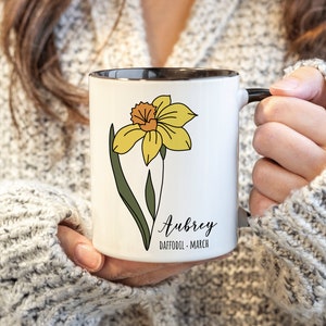 Peut inclure: Une tasse en c&eacute;ramique blanche avec un bord et une poign&eacute;e noirs. La tasse pr&eacute;sente une fleur de jonquille jaune avec des feuilles vertes et le texte "Aubrey Daffodil - March".