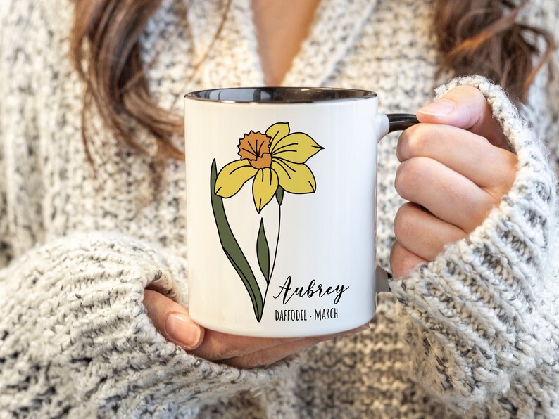 Peut inclure: Une tasse en c&eacute;ramique blanche avec un bord et une poign&eacute;e noirs. La tasse pr&eacute;sente une fleur de jonquille jaune avec des feuilles vertes et le texte "Aubrey Daffodil - March".