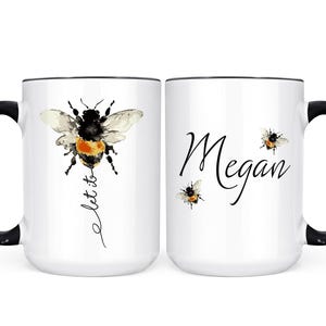 May include: White ceramic mug with a black handle and rim. One side features a watercolor bumblebee with the words "let it" in script. The other side has the name "Megan" in script with two smaller bee illustrations.