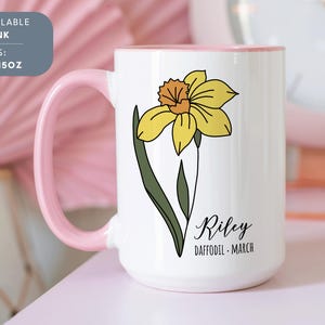 Peut inclure: Une tasse en c&eacute;ramique blanche avec un bord et une poign&eacute;e roses. La tasse pr&eacute;sente une illustration de jonquille jaune et le texte "Riley Daffodil - March".