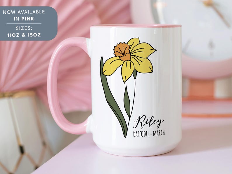 Peut inclure: Une tasse en c&eacute;ramique blanche avec un bord et une poign&eacute;e roses. La tasse pr&eacute;sente une illustration de jonquille jaune et le texte "Riley Daffodil - March".