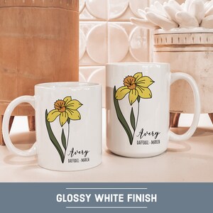 Peut inclure: Deux tasses en c&eacute;ramique blanche avec un motif de jonquille et le nom "Avery" avec le texte "Jonquille - Mars". Les tasses sont sur un comptoir en carrelage blanc.