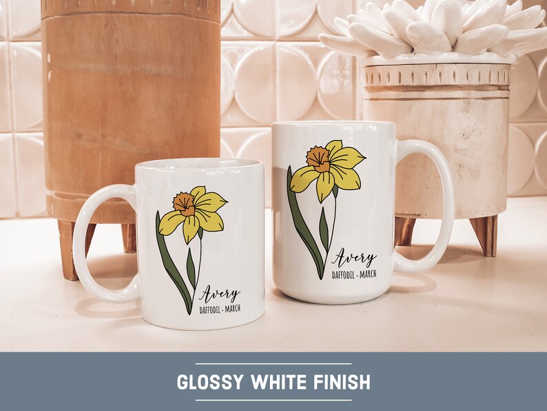Peut inclure: Deux tasses en c&eacute;ramique blanche avec un motif de jonquille et le nom "Avery" avec le texte "Jonquille - Mars". Les tasses sont sur un comptoir en carrelage blanc.