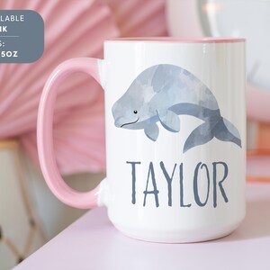 May include: White ceramic mug with a pink rim and handle. The mug features a watercolor illustration of a beluga whale and the name "TAYLOR" printed in black. The mug is available in 11oz and 15oz sizes.