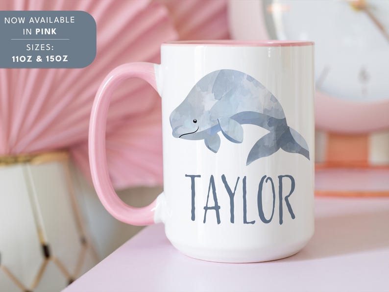May include: White ceramic mug with a pink rim and handle. The mug features a watercolor illustration of a beluga whale and the name "TAYLOR" printed in black. The mug is available in 11oz and 15oz sizes.