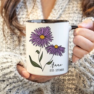 Puede incluir: Taza de cer&aacute;mica blanca con borde y asa negros. La taza presenta una ilustraci&oacute;n de una flor de aster p&uacute;rpura y el texto "Anne Aster - Septiembre".