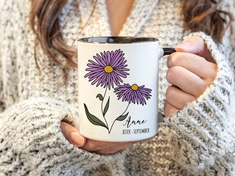 Puede incluir: Taza de cer&aacute;mica blanca con borde y asa negros. La taza presenta una ilustraci&oacute;n de una flor de aster p&uacute;rpura y el texto "Anne Aster - Septiembre".