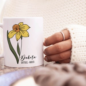Peut inclure: Une tasse en c&eacute;ramique blanche avec une illustration de jonquille jaune et le texte "Dakota Daffodil - March".