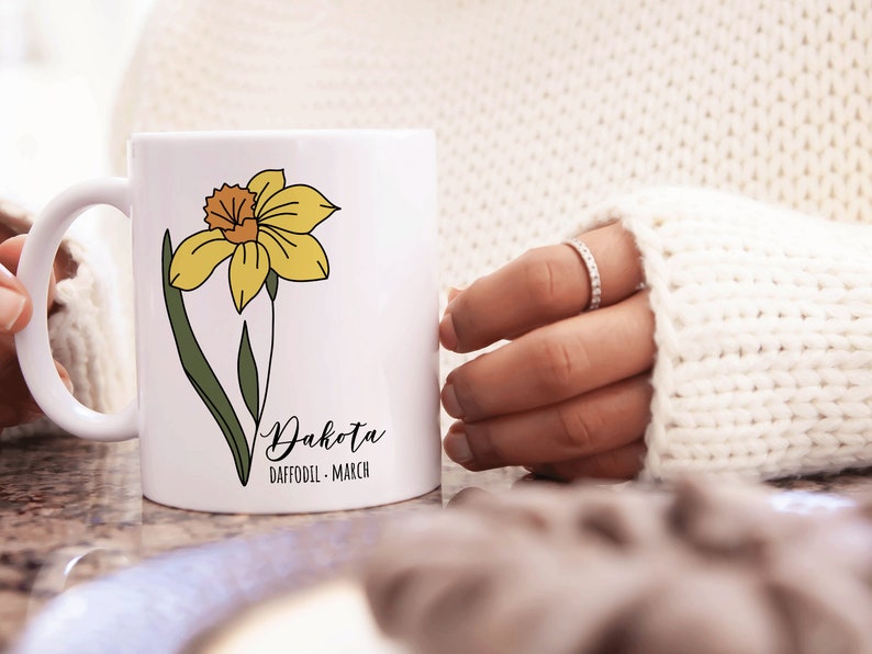 Peut inclure: Une tasse en c&eacute;ramique blanche avec une illustration de jonquille jaune et le texte "Dakota Daffodil - March".