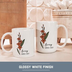 May include: Two white ceramic mugs with a floral design and the name "Avery" with the text "Gladiolus - August". The mugs are on a white countertop with a wooden bowl and a white ceramic planter in the background.
