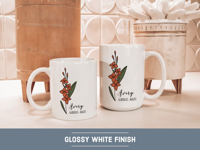 May include: Two white ceramic mugs with a floral design and the name "Avery" with the text "Gladiolus - August". The mugs are on a white countertop with a wooden bowl and a white ceramic planter in the background.