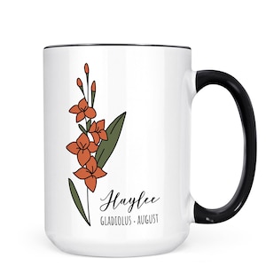 May include: White ceramic mug with a black handle. The mug has a black rim and features a line drawing of a gladiolus flower in orange and green. The text "Haylee Gladiolus - August" is printed on the mug.