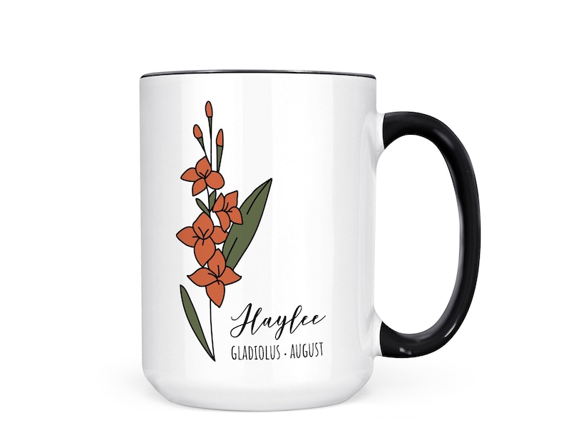May include: White ceramic mug with a black handle. The mug has a black rim and features a line drawing of a gladiolus flower in orange and green. The text "Haylee Gladiolus - August" is printed on the mug.