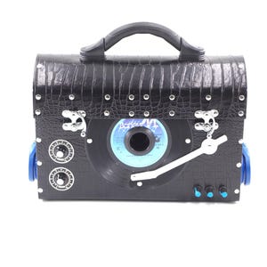 May include: A black leather record player case with a silver handle, a silver record player, and a silver tone arm. The case has silver studs and a blue and silver knob on the side.