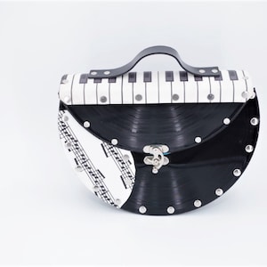 Könnte beinhalten: Eine schwarze und weiße Vinyl-Platten-Handtasche mit einem Klaviertasten-Design auf der oberen Klappe. Die Tasche hat einen schwarzen Griff und silberne Nieten an den Rändern.