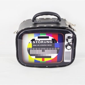 Könnte beinhalten: Eine schwarze Kunstleder-Handtasche in Form eines Vintage-Fernsehers mit einer durchsichtigen Plastikfront. Die Frontplatte zeigt ein buntes Fernseh-Testbild mit dem Text "STÖRUNG Aber wir arbeiten daran".