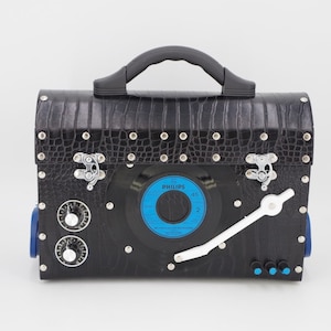 May include: A black leather briefcase with a vinyl record on the front. The briefcase has a handle, two silver clasps, and a white record player arm. The record is a 45 RPM record by Philips.