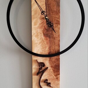 May include: A wooden clock with a black circular frame and Arabic calligraphy on the face. The clock is mounted on a wall.