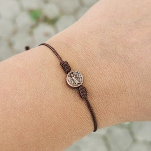 May include: A brown cord bracelet with a small, round, metal charm featuring a religious figure.