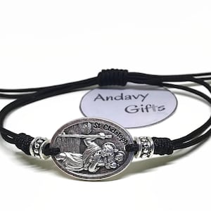 Puede incluir: Pulsera de cordón negro con un colgante ovalado plateado que representa a San Cristóbal. El colgante lleva la inscripción "St. Christopher". La pulsera tiene nudos ajustables y cuentas plateadas.