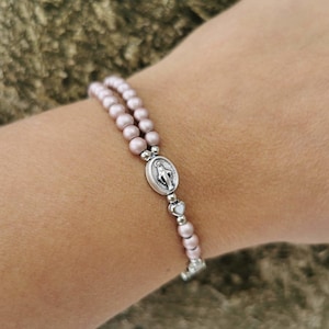 May include: A double-strand bracelet with pale pink beads and a silver-tone religious pendant. The bracelet is secured with a silver clasp. The beads are approximately 6 mm in diameter.