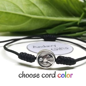 Puede incluir: Una pulsera de cordón negro con un dije circular plateado que presenta un diseño detallado de ángel. La pulsera tiene nudos ajustables y una pequeña cuenta plateada. El texto en la imagen dice "choose cord color".