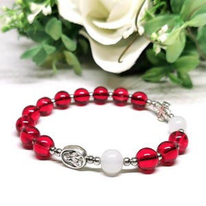 Può includere: Un braccialetto di perline rosse con una croce e un medaglione religioso color argento. Il braccialetto presenta perline rosse traslucide e perline bianche. Il braccialetto ha una circonferenza di circa 18 cm.