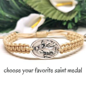 May include: Beige macrame bracelet with an oval silver saint medal. The bracelet has a sliding knot closure with silver beads. The text "choose your favorite saint medal" is below the bracelet.