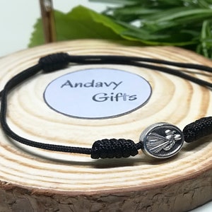 May include: Black cord bracelet with a silver oval charm featuring a depiction of the Virgin Mary. The bracelet is tied with a knot and has silver beads on the ends. The charm is centered on the bracelet and is the focal point of the image. The bracelet is displayed on a wooden surface.