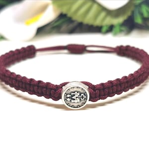 May include: Maroon macrame bracelet with a silver-toned centerpiece. The bracelet features a woven design and adjustable sliding knot closure. The centerpiece has an embossed religious symbol.