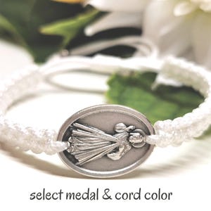 May include: White macrame bracelet featuring a silver oval pendant with a religious figure. The bracelet has a woven cord design. Text at the bottom reads "select medal & cord color".