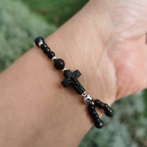 Puede incluir: Pulsera de cuentas negras con un colgante de crucifijo. La pulsera tiene pequeñas cuentas redondas negras y detalles plateados. El colgante de la cruz también es negro y tiene un diseño detallado.