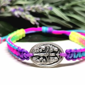 May include: A colorful macrame bracelet with a silver oval charm. The bracelet features a vibrant mix of pink, blue, yellow, and purple threads. The charm has a detailed religious design.