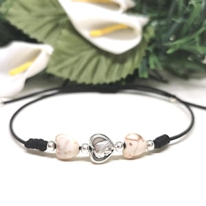 Puede incluir: Pulsera ajustable de cordón negro con dos piedras claras en forma de corazón y un dije plateado en forma de corazón. La pulsera tiene cuentas plateadas y se exhibe sobre un fondo de flores blancas y follaje verde.