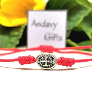 Könnte beinhalten: Ein rotes Kordelarmband mit einem silberfarbenen ovalen Anhänger. Der Anhänger hat ein detailliertes Design mit einem Kreuz und anderen Symbolen. Das Armband ist auf beiden Seiten des Anhängers geknotet. Der Hintergrund enthält ein verschwommenes Schild mit "Andavy Gifts".