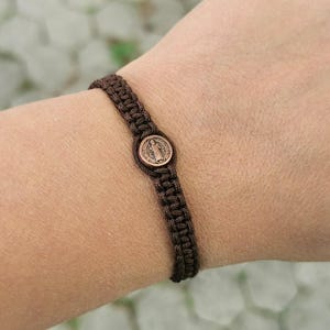 May include: A brown macramé bracelet with a bronze medallion depicting a religious figure. The bracelet, made of woven cord, is worn on a wrist. The circular medallion has a detailed design.
