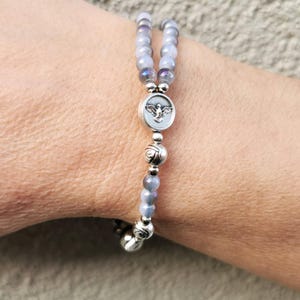 May include: A beaded bracelet featuring light blue and purple beads. The bracelet includes a silver charm with an angel design and decorative silver beads. The bracelet is worn on a wrist.