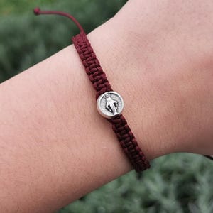 Puede incluir: Una pulsera de macramé burdeos con un medallón plateado que representa una figura religiosa. La pulsera ajustable está anudada y tiene un cierre de nudo corredizo. La pulsera se lleva en una muñeca.
