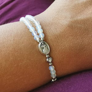 May include: A white beaded bracelet with a silver charm featuring a religious figure. The bracelet is made of small, round, opalescent beads and is worn on a wrist. The background is a purple fabric.