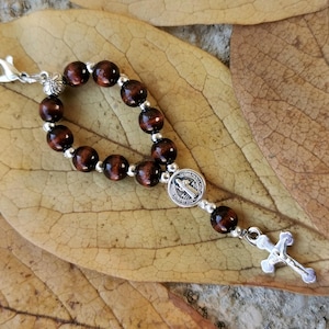 May include: A small rosary with dark brown wooden beads and silver-coloured accents. The rosary features a cross pendant and a medallion with a religious figure. The rosary is displayed on a dried leaf.