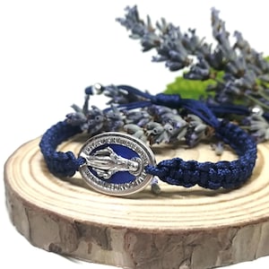 Può includere: Un braccialetto in macramè blu navy con un ciondolo ovale argentato raffigurante un'immagine religiosa. Il braccialetto è esposto su una fetta di legno con rametti di lavanda sullo sfondo. Il ciondolo ha smalto blu.