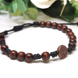 Puede incluir: Una pulsera con cuentas de madera de color marrón oscuro ensartadas en un cordón negro con nudos ajustables. Un dije circular de metal cobrizo con una figura religiosa en relieve está integrado en la secuencia de cuentas.