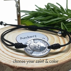 Könnte beinhalten: Schwarzes Kordelarmband mit einem silberfarbenen ovalen Anhänger mit einem Heiligenbild. Das Armband hat verstellbare Knoten und eine kleine silberne Perle. Auf dem Anhänger steht "ST. CHARBEL". Der Text "choose your saint & color" ist ebenfalls sichtbar.