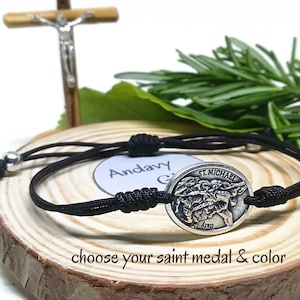 May include: A black cord bracelet with a silver St. Michael medal. The bracelet is displayed on a wooden surface with a small wooden crucifix and greenery in the background. Text on the image reads "choose your saint medal & color."