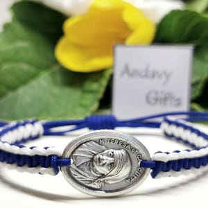 Könnte beinhalten: Ein blau-weiß geflochtenes Armband mit einem silbernen ovalen Anhänger mit einem Bild von Mutter Teresa und dem Text "M. TERESA OF CALCUTTA". Das Armband hat einen blauen Kordelverschluss und ist vor einem unscharfen Hintergrund platziert.